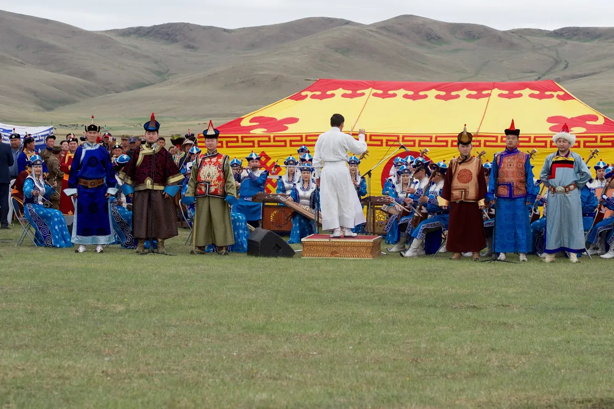 A conductor in white faces an orchestra in blue costume, with soloists in brocade robes standing beside it, in front of a red and yellow tent on the steppe near Ulaanbaatar